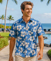Man wearing a blue button up Aloha Friday floral shirt standing in a tropical setting with palm trees and ocean.
