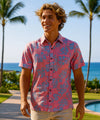 Man wearing a button up floral coral Aloha shirt standing in a tropical setting with palm trees and ocean.