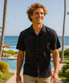 Man in a a Hawaiian aloha black shirt standing by a pool with palm trees and ocean in the background