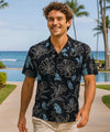 Man wearing a black floral rayon poplin shirt standing in front of a tropical beach with palm trees.