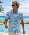Man wearing a blue patterned shirt and sunglasses on a beach with palm trees.